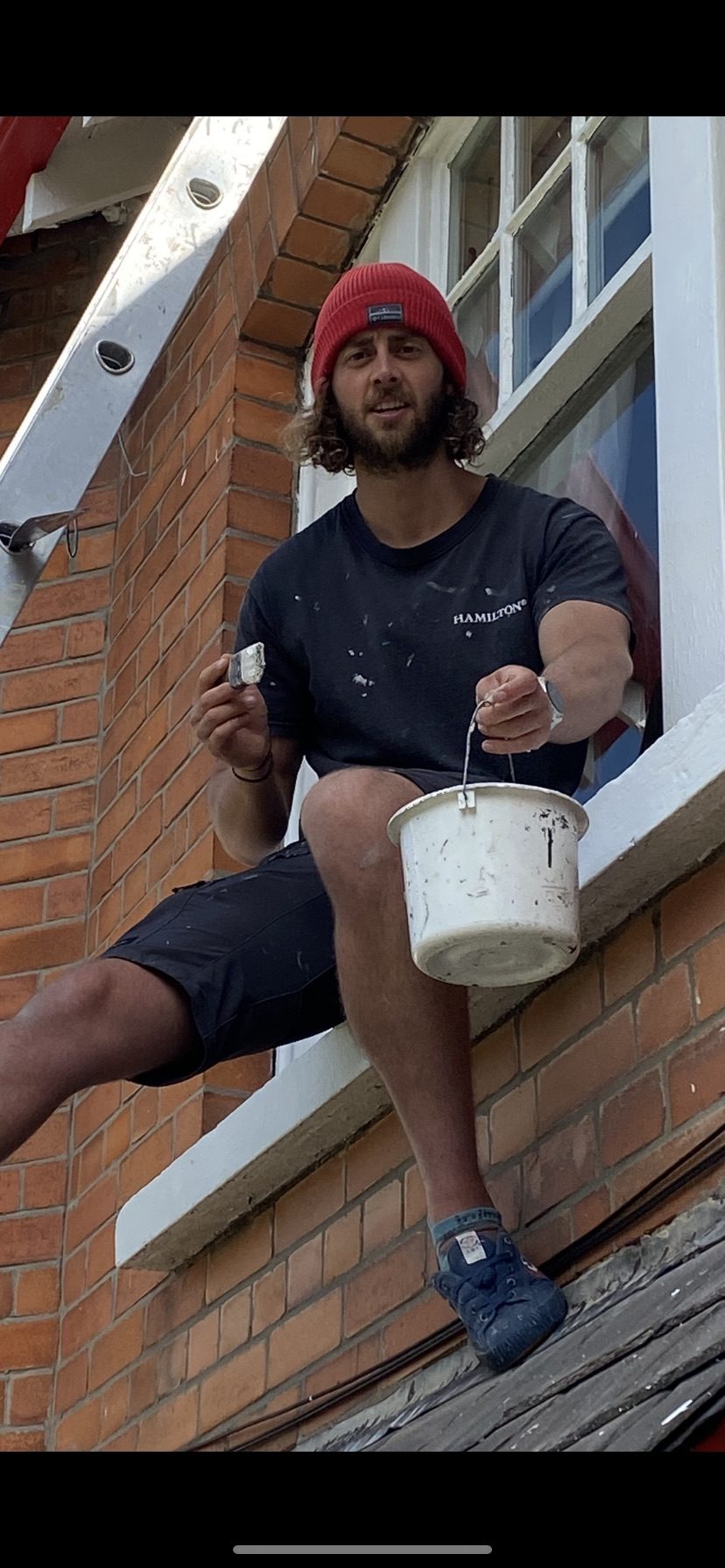 Caolán sitting on a window ledge holding a paint bucket, in front of a red-brick wall